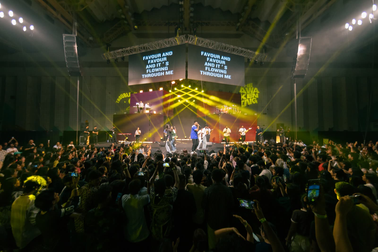 A packed room worshipping toward a brightly lit stage with lyrics on the screens